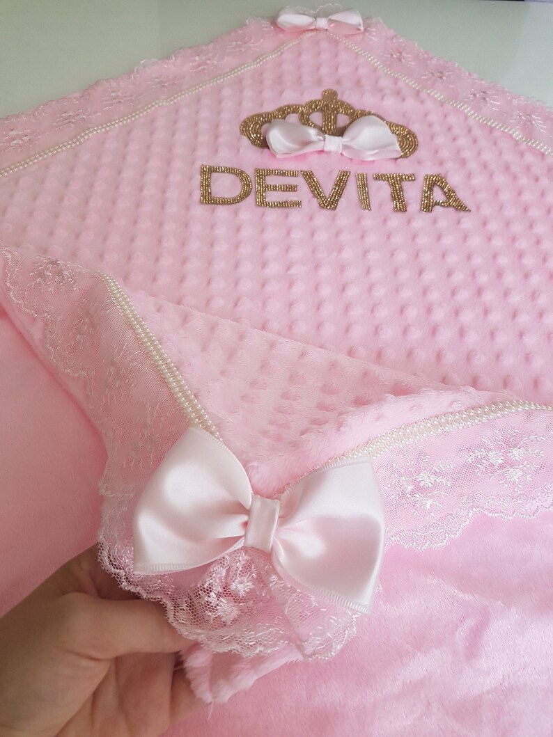 May include: A pink baby blanket with a white lace trim and a gold crown with the name "DEVITA" embroidered on it.