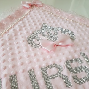 May include: Pink baby blanket with a silver crown and the word "GIRL" spelled out in silver rhinestones. The blanket has a pearl trim and a pink bow.