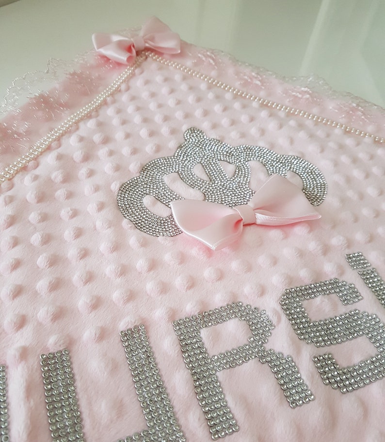 May include: Pink baby blanket with a silver crown and the word "GIRL" spelled out in silver rhinestones. The blanket has a pearl trim and a pink bow.