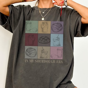 May include: A dark gray t-shirt with a graphic of various bread and jar items in a grid pattern. The text "IN MY SOURDOUGH ERA" is printed below the graphic.