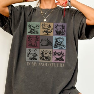 Könnte beinhalten: Ein dunkelgraues T-Shirt mit einer Grafik von neun Axolotln in verschiedenen Posen. Die Axolotl sind in einem Raster angeordnet und jeder hat einen anderen Ausdruck. Der Text "In My Axolotl Era" ist unter dem Raster gedruckt.
