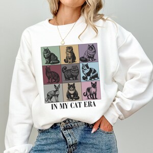 K&ouml;nnte beinhalten: Wei&szlig;er Pullover mit einem Grafikdruck von neun verschiedenen Katzen in bunten Quadraten. Der Text "IN MY CAT ERA" ist unter dem Grafikdruck aufgedruckt.