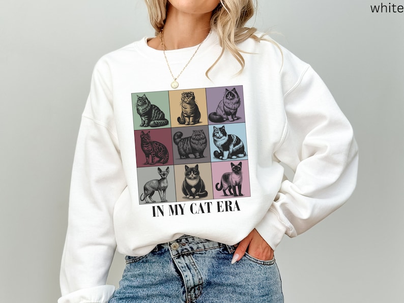 K&ouml;nnte beinhalten: Wei&szlig;er Pullover mit einem Grafikdruck von neun verschiedenen Katzen in bunten Quadraten. Der Text "IN MY CAT ERA" ist unter dem Grafikdruck aufgedruckt.