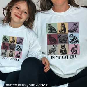 K&ouml;nnte beinhalten: Zwei wei&szlig;e Sweatshirts mit Rundhalsausschnitt und einem Motiv aus neun verschiedenen Katzen in bunten Quadraten. Der Text "In My Cat Era" ist unter dem Motiv aufgedruckt. Die Sweatshirts werden von einer Frau und einem Kind getragen.