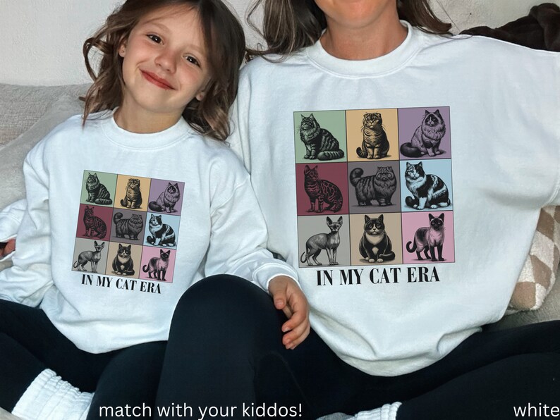 K&ouml;nnte beinhalten: Zwei wei&szlig;e Sweatshirts mit Rundhalsausschnitt und einem Motiv aus neun verschiedenen Katzen in bunten Quadraten. Der Text "In My Cat Era" ist unter dem Motiv aufgedruckt. Die Sweatshirts werden von einer Frau und einem Kind getragen.