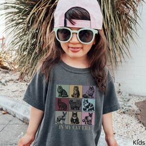 K&ouml;nnte beinhalten: Ein Kind tr&auml;gt ein graues T-Shirt mit einer Grafik von neun Katzen in bunten Quadraten. Das Shirt sagt "In My Cat Era". Das Kind tr&auml;gt auch eine rosa Baseballkappe und eine gr&uuml;ne Sonnenbrille.