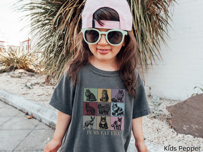 K&ouml;nnte beinhalten: Ein Kind tr&auml;gt ein graues T-Shirt mit einer Grafik von neun Katzen in bunten Quadraten. Das Shirt sagt "In My Cat Era". Das Kind tr&auml;gt auch eine rosa Baseballkappe und eine gr&uuml;ne Sonnenbrille.