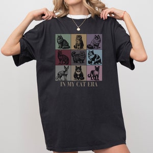 K&ouml;nnte beinhalten: Schwarzes T-Shirt mit einer Grafik von neun verschiedenen Katzen in bunten Quadraten. Der Text "In My Cat Era" ist unter der Grafik gedruckt.