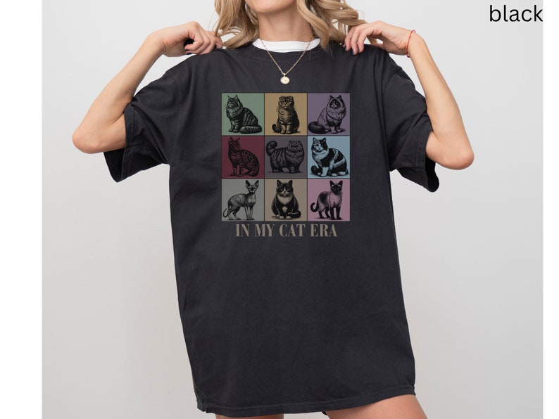 K&ouml;nnte beinhalten: Schwarzes T-Shirt mit einer Grafik von neun verschiedenen Katzen in bunten Quadraten. Der Text "In My Cat Era" ist unter der Grafik gedruckt.