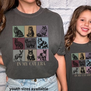 K&ouml;nnte beinhalten: Zwei Personen tragen anthrazitfarbene T-Shirts mit einer Vintage-Grafik von neun Katzen in bunten Quadraten. Der Text "In My Cat Era" ist unter der Grafik gedruckt. Die Person links tr&auml;gt Jeans und die Person rechts tr&auml;gt ein anthrazitfarbenes T-Shirt. Der Text "youth sizes available" ist unter dem Bild gedruckt.