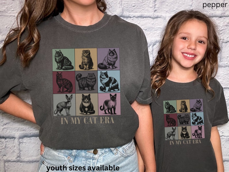 K&ouml;nnte beinhalten: Zwei Personen tragen anthrazitfarbene T-Shirts mit einer Vintage-Grafik von neun Katzen in bunten Quadraten. Der Text "In My Cat Era" ist unter der Grafik gedruckt. Die Person links tr&auml;gt Jeans und die Person rechts tr&auml;gt ein anthrazitfarbenes T-Shirt. Der Text "youth sizes available" ist unter dem Bild gedruckt.