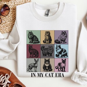 K&ouml;nnte beinhalten: Wei&szlig;er Pullover mit einer Grafik von neun verschiedenen Katzen in einem Rastermuster. Der Text "IN MY CAT ERA" ist unter dem Raster gedruckt.