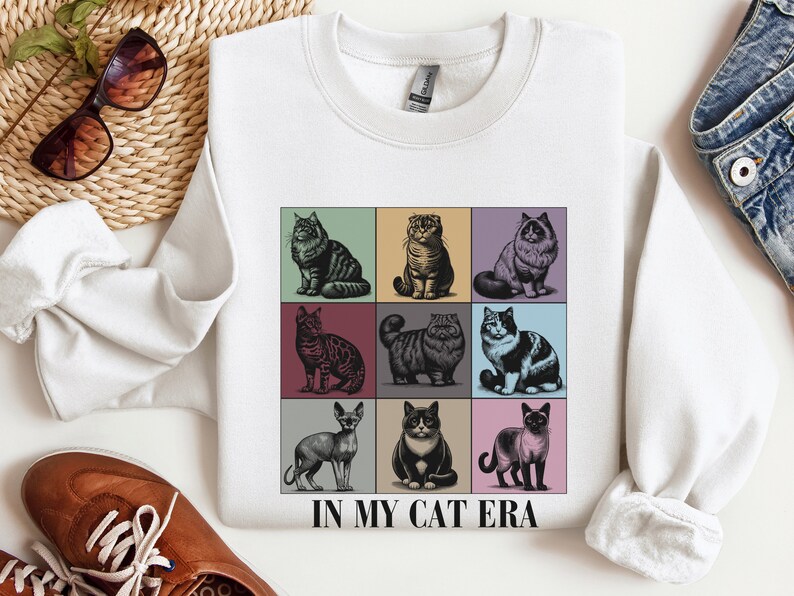K&ouml;nnte beinhalten: Wei&szlig;er Pullover mit einer Grafik von neun verschiedenen Katzen in einem Rastermuster. Der Text "IN MY CAT ERA" ist unter dem Raster gedruckt.