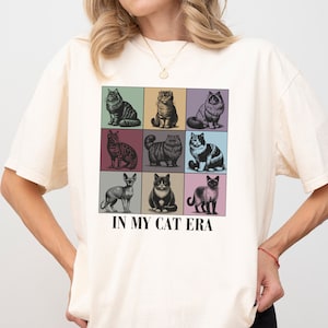 K&ouml;nnte beinhalten: Ein wei&szlig;es T-Shirt mit einer Grafik von neun Katzen in bunten Quadraten. Der Text "IN MY CAT ERA" ist unter der Grafik aufgedruckt. Das Shirt ist in Elfenbein erh&auml;ltlich.