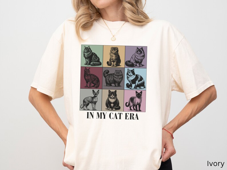 K&ouml;nnte beinhalten: Ein wei&szlig;es T-Shirt mit einer Grafik von neun Katzen in bunten Quadraten. Der Text "IN MY CAT ERA" ist unter der Grafik aufgedruckt. Das Shirt ist in Elfenbein erh&auml;ltlich.