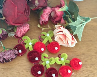 May include: A collection of red and burgundy artificial flowers and cherry ornaments with green stems and bows. The cherries have white accents. A decorative cup with cat illustrations is in the background.