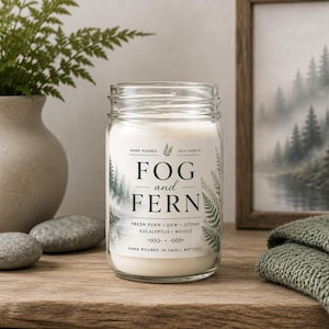 May include: A clear glass jar candle filled with white wax. The label features the text FOG and FERN, along with the scent notes fresh fern, dew, ozone, eucalyptus, and woods. The design includes illustrations of pine trees and fern fronds.