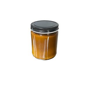 May include: A clear glass jar filled with a golden-brown substance, topped with a black lid. The jar is cylindrical and appears to be filled to the top. The background is plain white.