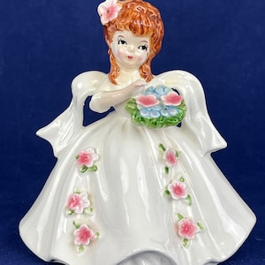 Może przedstawiać: Figurka z porcelany w stylu vintage przedstawiająca kobietę w białej sukni, ozdobioną różowymi kwiatowymi detalami. Figurka ma rude włosy i trzyma bukiet niebieskich i różowych kwiatów. Różowy kwiat jest we włosach.