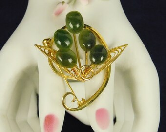 May include: A vintage gold-tone brooch featuring five oval, green jade stones arranged in a stylized floral design. The brooch has intricate filigree detailing and a curved pin closure, showcasing a classic, elegant aesthetic.