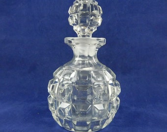 May include: A clear, faceted glass perfume bottle with a matching stopper. The bottle has a rounded shape with a geometric pattern and a flared neck. The stopper is also faceted and has a decorative, geometric design. The bottle is empty and sits against a blue background.