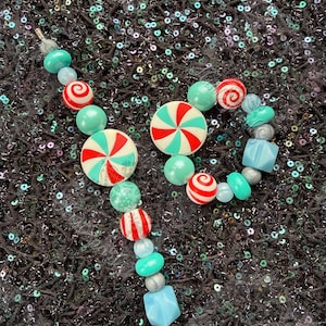 May include: A beaded necklace and bracelet set featuring a candy theme. The jewelry includes round beads in teal, red, white, and silver, with larger candy-shaped pendants. The background is a dark, textured surface with iridescent sequins.