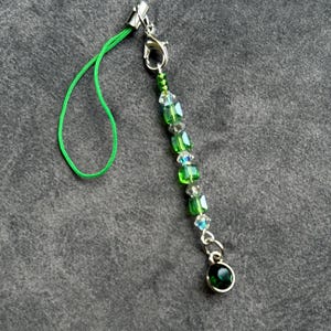 May include: A green and silver beaded phone charm. The charm features a green cord, a silver clasp, and a strand of green and clear beads. A small, round, green gem hangs from the bottom.