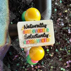 May include: A white rectangular charm with the words "Naturally Introverted Selectively Extroverted" in colorful lettering. The charm is adorned with yellow and green spherical accents. The background is a dark, glittery surface.