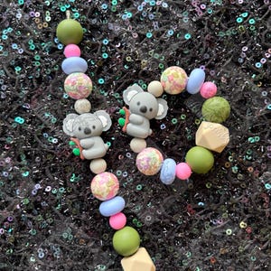 May include: A beaded koala bear-themed accessory featuring gray koala charms, floral and pastel-colored beads, and geometric shapes. The beads are strung together, creating a playful and colorful design.