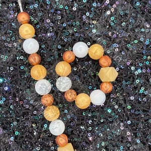 May include: A beaded necklace with a variety of beads in shades of gold, white, and brown. The beads are round, with some featuring a patterned design. The necklace is displayed on a black, sequined surface.