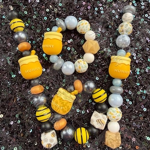 May include: A collection of colorful beads arranged in a decorative pattern. The beads include yellow and black striped spheres, honey pot charms with the word "HUNNY", and various other shapes and colors. The background is a dark, sequined fabric.