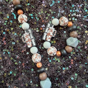 May include: Two beaded accessories featuring round silicone beads in brown, black, white, and gray, along with faceted gray beads. Each accessory includes a flat, rectangular charm shaped like a tumbler cup with the text MAMA'S SIPPY CUP.