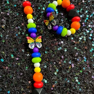 May include: Two colorful beaded bracelets. One is a strand of rainbow-colored beads with a butterfly charm and a black geometric bead. The other is a circular bracelet with rainbow beads, a butterfly charm, and a black geometric bead.