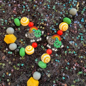 May include: A beaded bracelet with a St. Patrick's Day theme. The bracelet features a variety of colorful beads, including green, yellow, red, and white. Two charms with a rainbow and a pot of gold are also included. The bracelet is set against a black, sequined background.