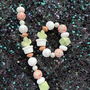 May include: A beaded teething necklace featuring a cactus theme. The necklace includes white, pink, and green beads, along with cactus-shaped charms in white pots. The beads have various patterns and shapes, creating a playful and decorative accessory.