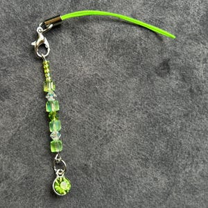May include: A green beaded charm with a lobster clasp and a lime green cord. The charm features a variety of green beads, including small round beads, square beads, and clear crystal beads. A small, round, green jewel hangs at the bottom.