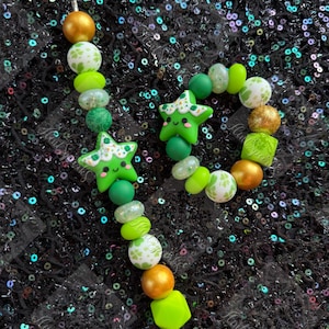 May include: A beaded jewelry set featuring green and gold beads. The set includes a necklace and a bracelet. The necklace has a star-shaped charm with a smiling face. The bracelet has a circular design with various bead shapes and sizes.