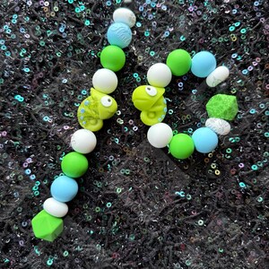May include: A pair of handmade beaded earrings featuring a playful design. The earrings include white, green, and blue beads, along with small, light green chameleon charms. The word "Fluff" is printed on a blue bead.