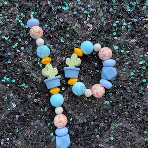 May include: A beaded keychain in the shape of the letter "P". The keychain features a variety of beads in shades of blue, white, orange, and pink. Some beads have cactus designs. The background is a dark, sequined fabric.