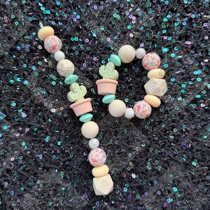 May include: A handmade beaded keychain featuring a cactus and floral theme. The keychain includes light pink, white, and turquoise beads, along with small cactus charms in pink pots. The design is arranged in a whimsical pattern.