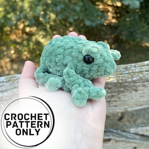 May include: A green crocheted frog amigurumi toy with black eyes. The frog is sitting in a person's hand. The image includes the text "CROCHET PATTERN ONLY".
