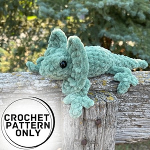 Op de afbeelding: Een groene, gehaakte kameleon met zwarte ogen zit op een verweerd houten hek. De kameleon heeft grote, gekrulde oren. De afbeelding bevat de tekst "CROCHET PATTERN ONLY".