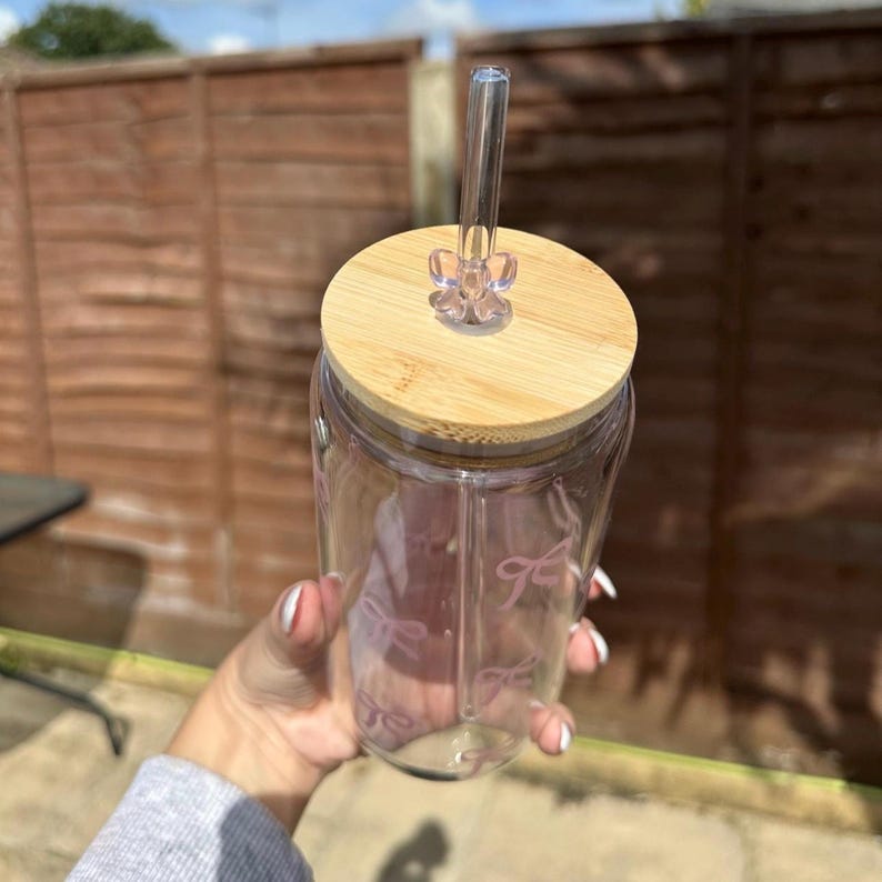 May include: A clear glass tumbler with a wooden lid and a clear straw. The tumbler has a pink bow pattern and is held by a person. The lid has a clear plastic stopper to hold the straw in place.