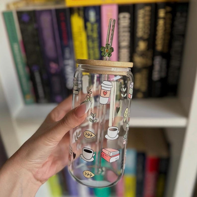 May include: A clear glass tumbler with a bamboo lid and a green flower-shaped straw topper. The tumbler is decorated with coffee-themed illustrations, including coffee cups, books, and leaves. The tumbler is held in front of a bookshelf.