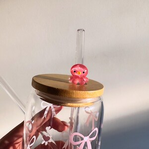 May include: A clear glass tumbler with a bamboo lid and a pink octopus charm. The glass is decorated with pink bows and hearts. A clear glass straw is inserted into the tumbler.