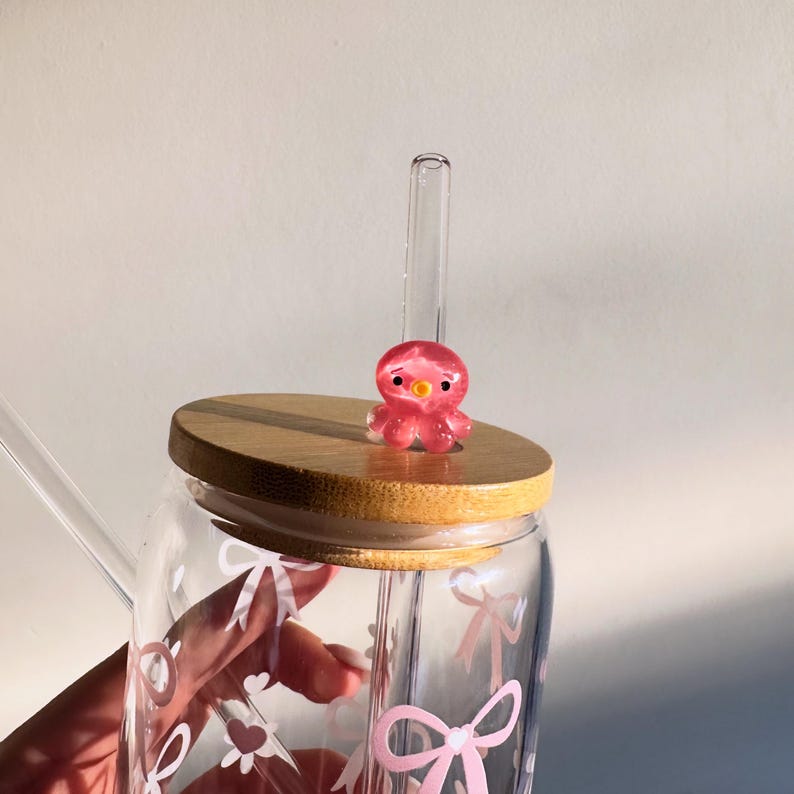 May include: A clear glass tumbler with a bamboo lid and a pink octopus charm. The glass is decorated with pink bows and hearts. A clear glass straw is inserted into the tumbler.