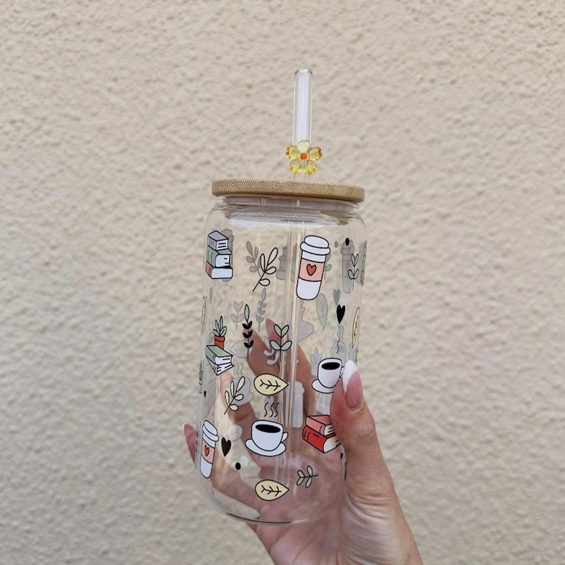 May include: A clear glass tumbler with a bamboo lid and a decorative straw. The tumbler is adorned with whimsical illustrations of books, coffee cups, and leaves. A small yellow flower decorates the top of the straw.