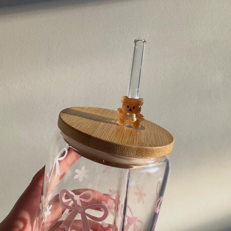 May include: A clear glass tumbler with a bamboo lid and a clear glass straw. The tumbler is decorated with pink bows and flowers. A small, brown teddy bear figurine sits on the lid.
