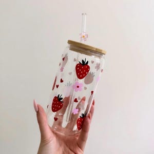 May include: A clear glass tumbler with a bamboo lid and a glass straw. The tumbler is decorated with red strawberries, pink flowers, and hearts. The straw has a small pink flower charm.