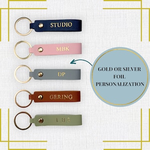 May include: Five leather keychains with gold lettering. The keychains are in different colors: navy blue, pink, gray, brown, and green. The lettering on the keychains reads: "STUDIO", "MBK", "DP", "GERING", and "L.B.S". A circle with a gold outline and a light blue background reads "GOLD OR SILVER FOIL PERSONALIZATION".